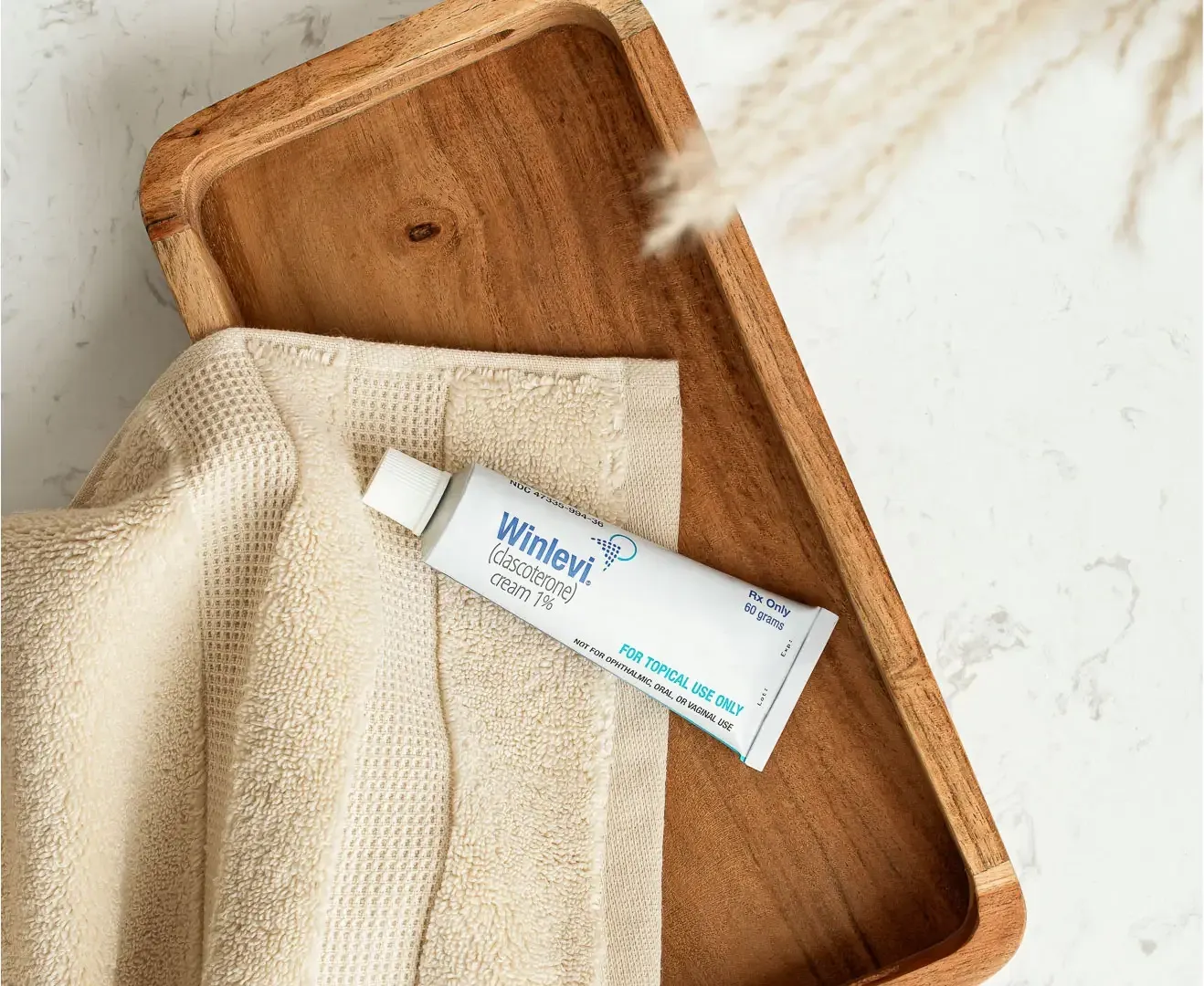 A Winlevi skincare tube resting on a folded cream towel in a wooden tray, shot on marble with a sprig of dried grass casting a soft shadow.