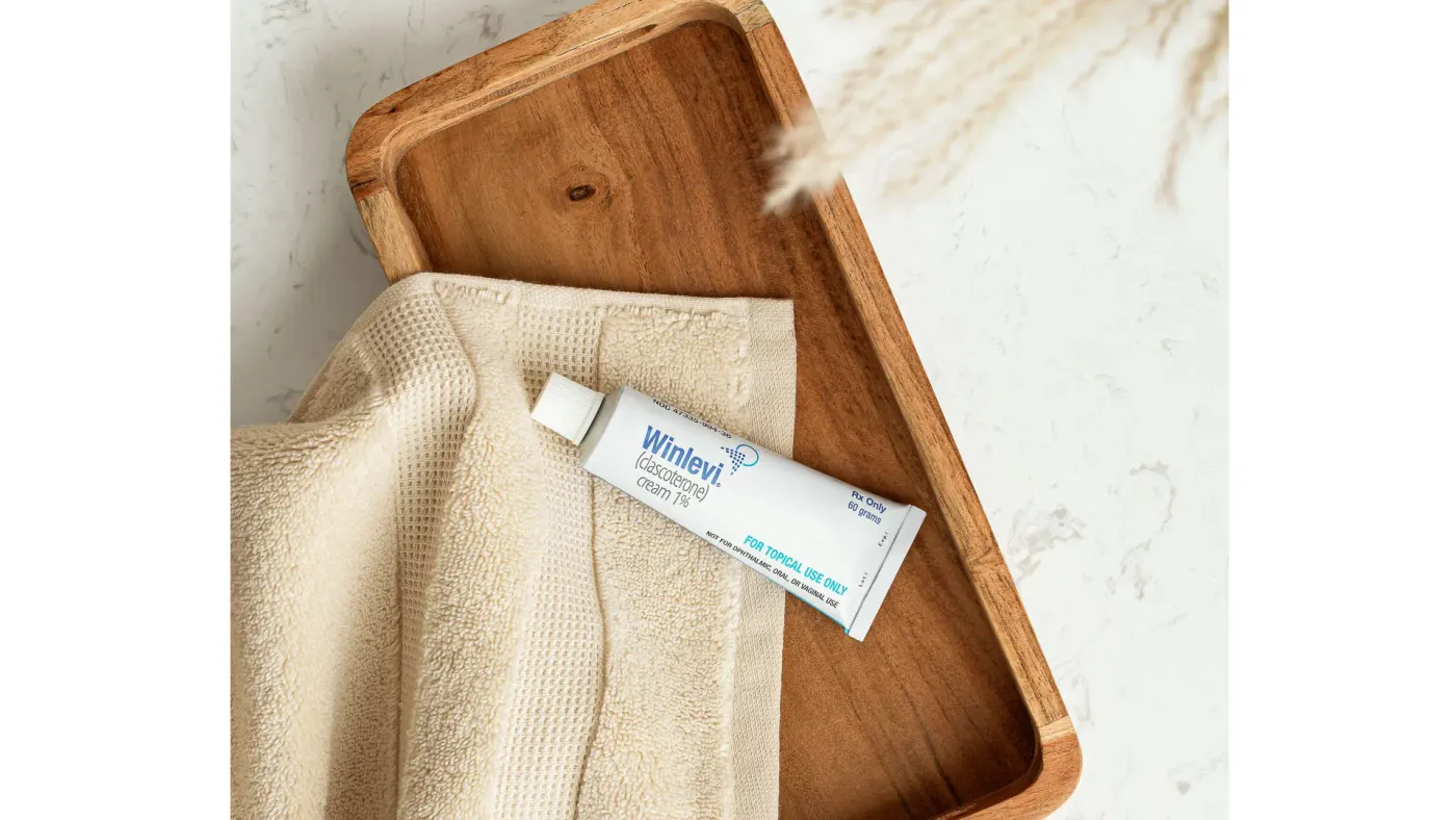 A Winlevi skincare tube resting on a folded cream towel in a wooden tray, shot on marble with a sprig of dried grass casting a soft shadow.