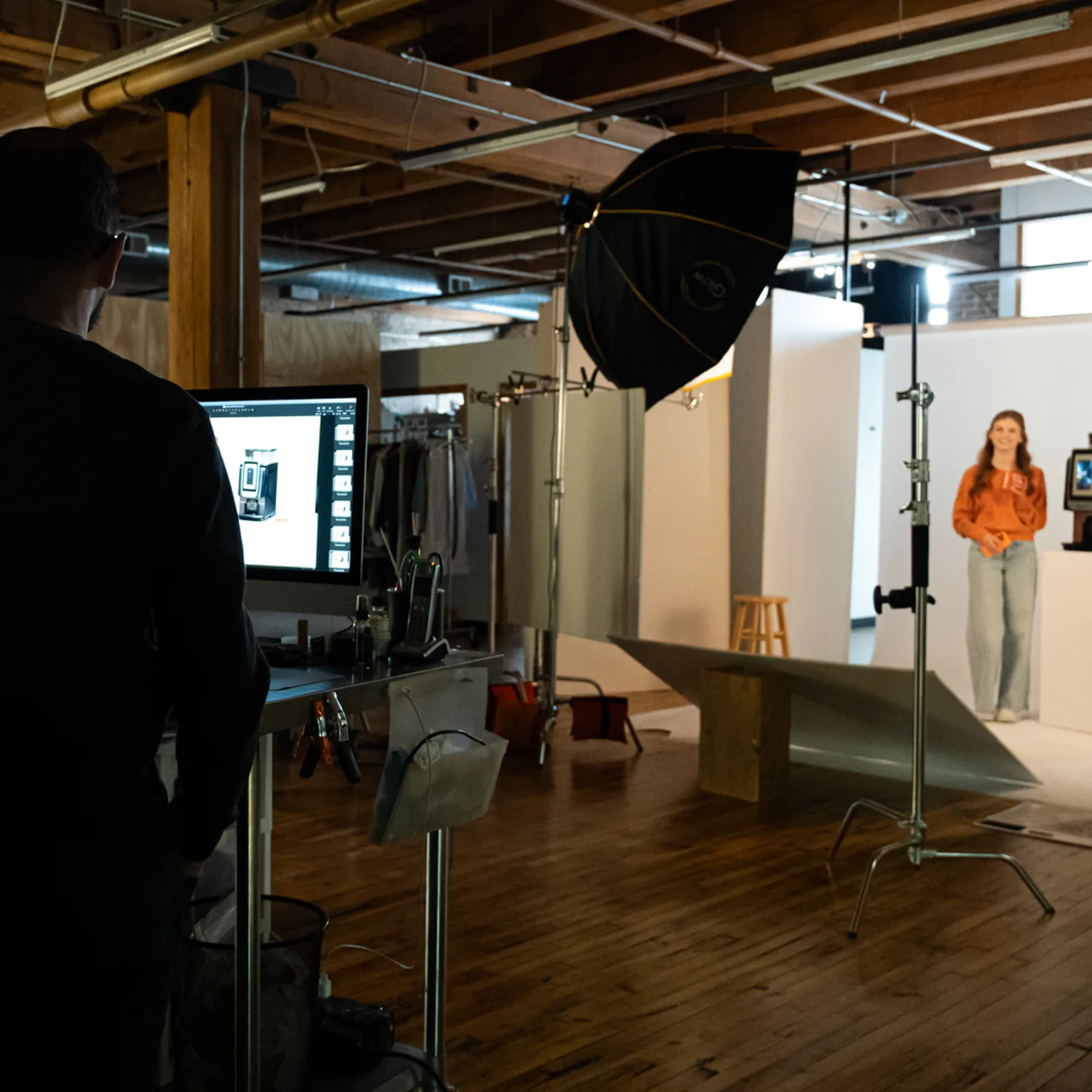 Behind the scenes of a WithMe product shoot: a crew member watches captured images on a monitor while a model stands on a white set under a large softbox